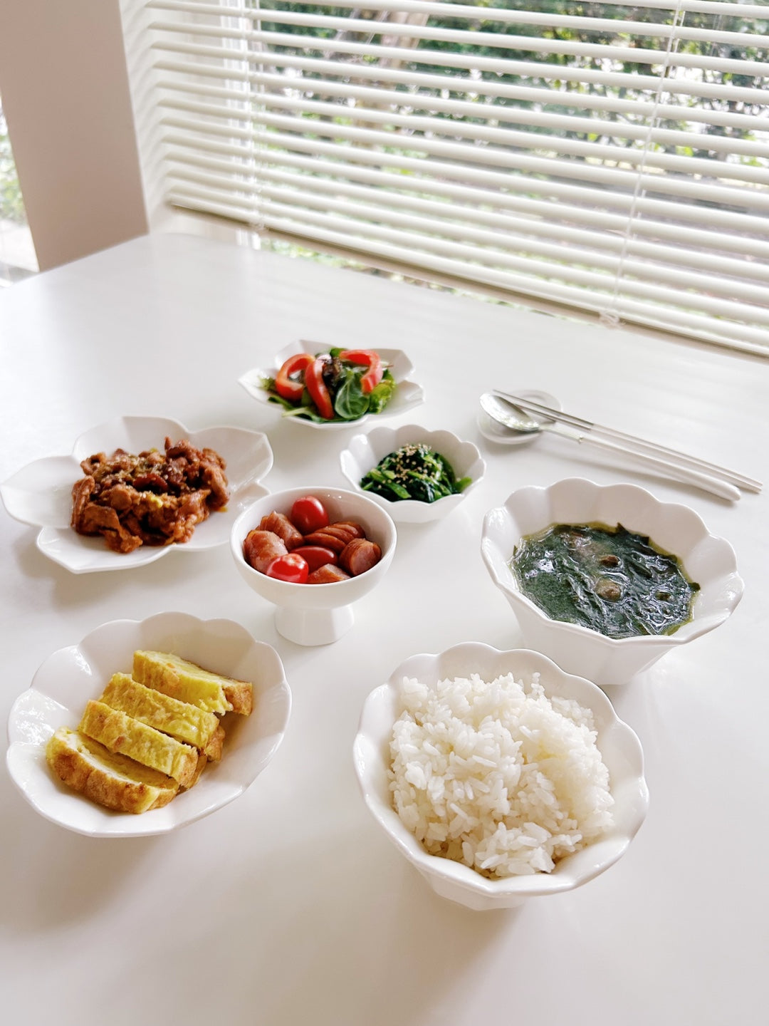 Assorted dishes including rice, vegetables, and meat on a white table with a window in the background.