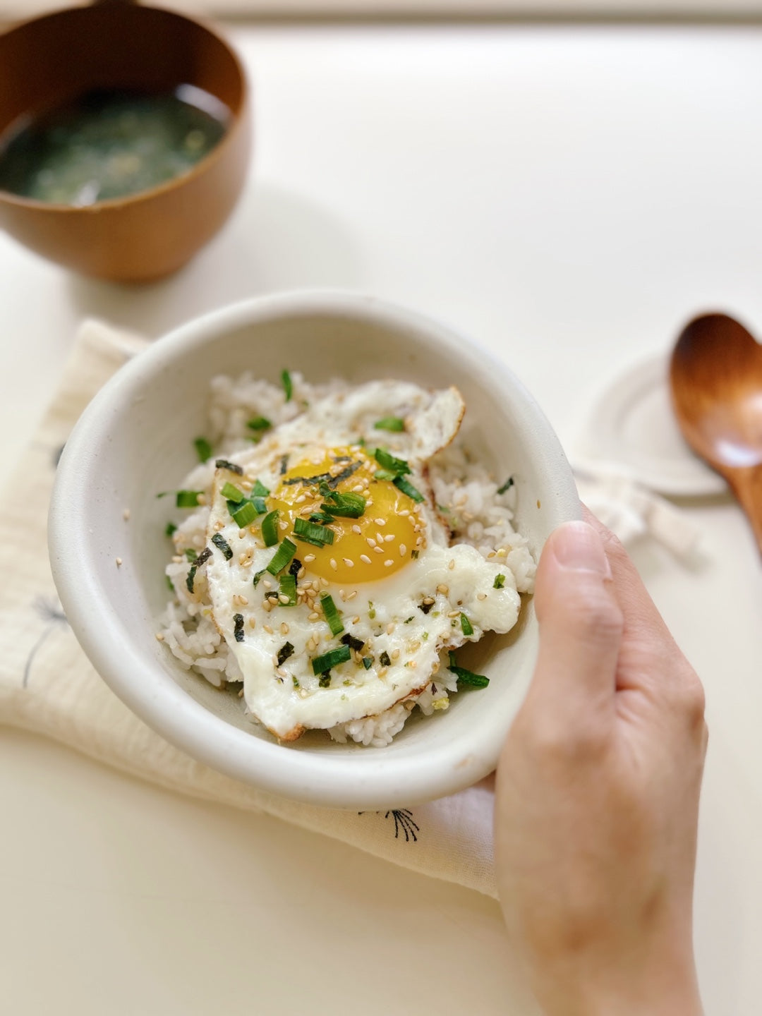 White bowl with a fried egg on rice, garnished with green onions, on a light surface.