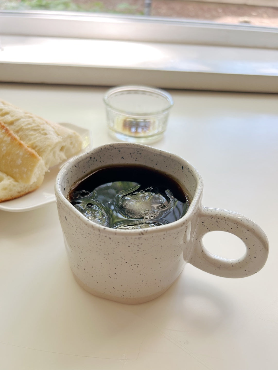 Ceramic mug with dark liquid and ice cubes on a light surface, with bread and a glass in the background.
