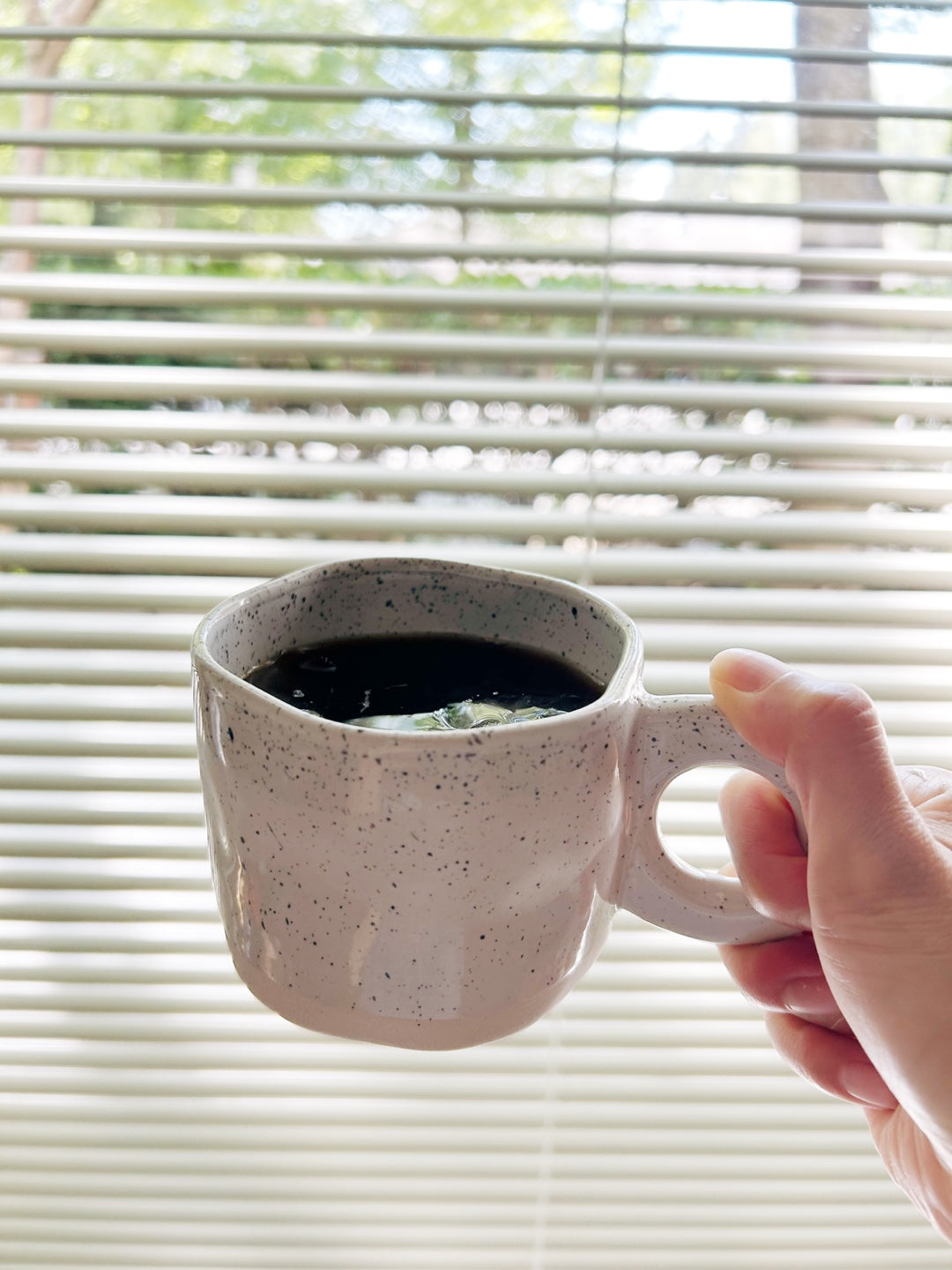 Person holding a speckled ceramic mug with a dark liquid in front of window blinds