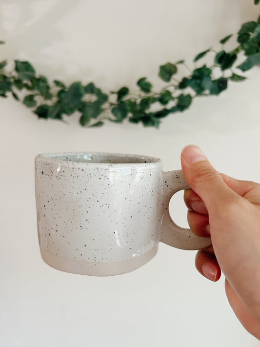 Hand holding a speckled ceramic mug with a white background and greenery.